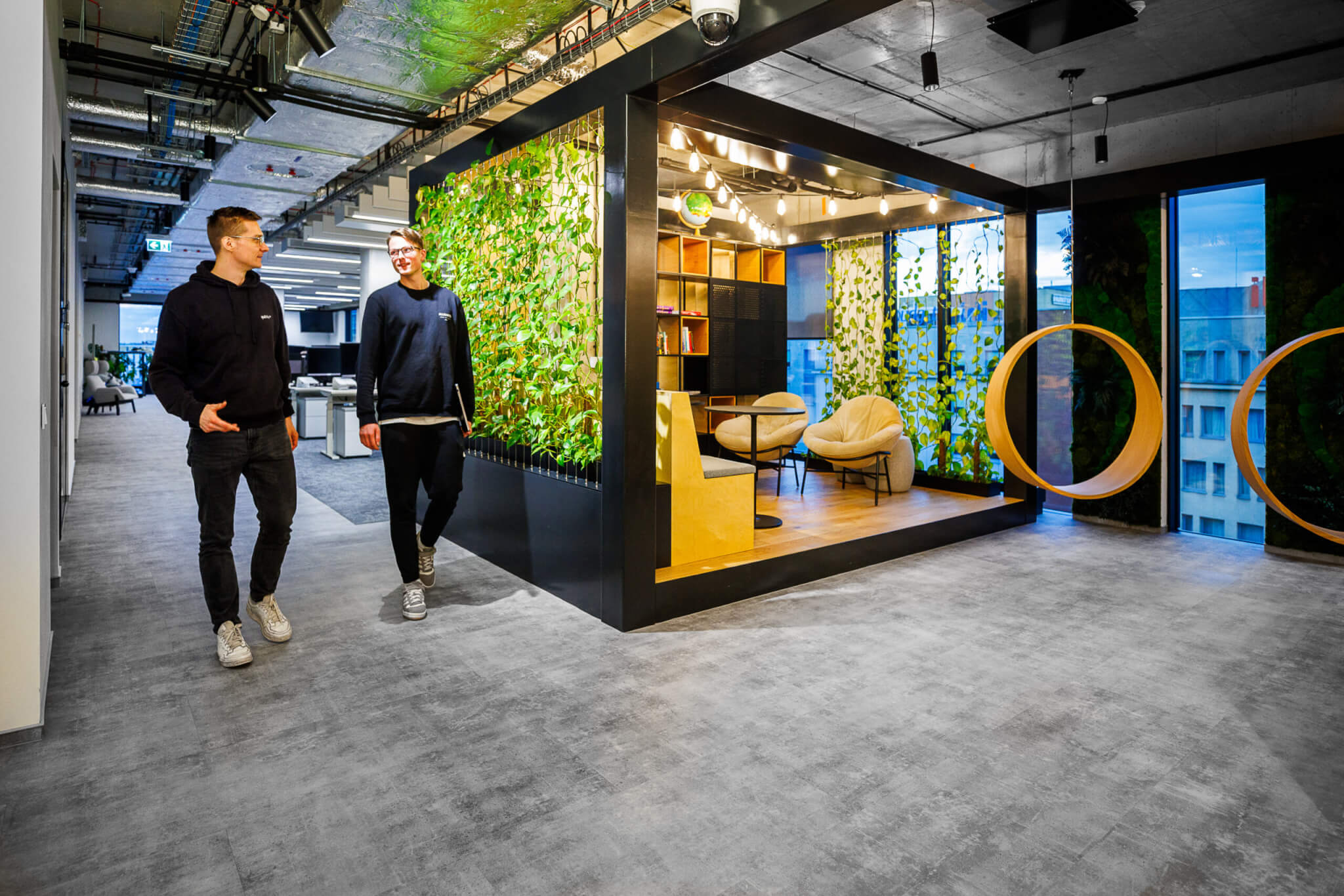 Qualtricians at the Krakow, Poland office during a walking meeting. They're walking down a hallway next to an indoor garden meeting space. The hallway is divided by a wall of greenery, with hanging chairs just next to the meeting space.