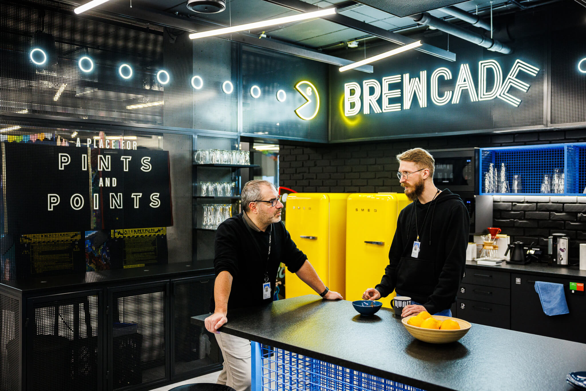 Two Qualtrics employees are in the Brewcade, the Krakow office coffee and lunch area, talking. There is are bright yellow refrigerators in the background and a sign that says 'a place for pints and points'.