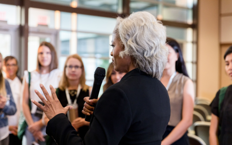 A woman speaking to an engaged audience at a professional event, holding a microphone and gesturing expressively.