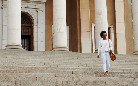 A woman walking down the steps of a large columned institutional building, smiling and carrying a shoulder bag.