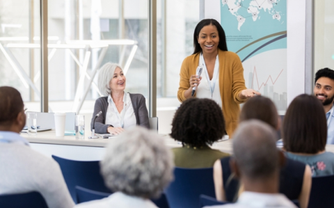 A woman presenting to a group of colleagues in a bright conference room, smiling and holding a pen.