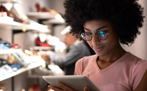 A retail store employee reviewing a tablet on the sales floor, with illuminated shoe displays in the background.