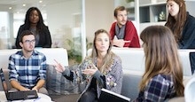 A group of people sitting and standing around a table, discussing ideas.