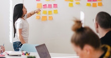 A group of people in a meeting room, one is looking at a computer, one is pointing to a white board covered in post it notes and one is looking at that whiteboard