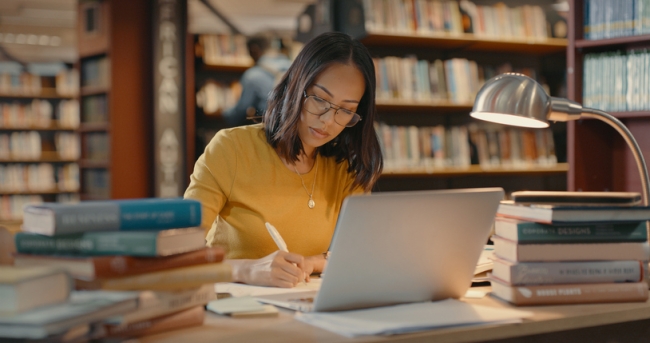 Woman conducting in-depth research on a laptop and with multiple books in a university library.