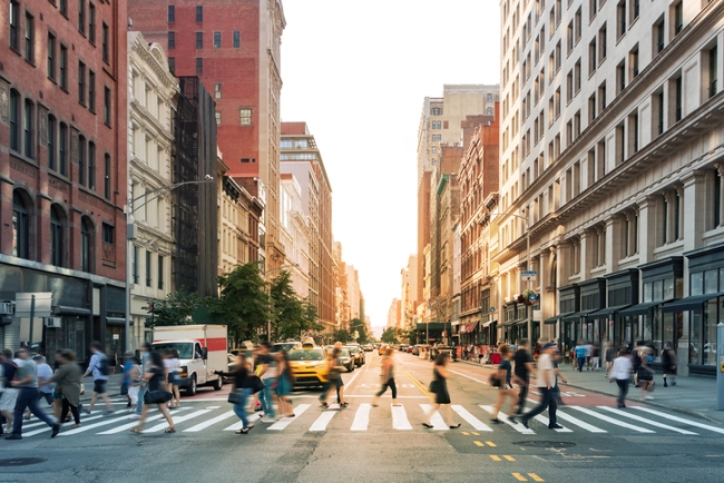 Street-level view of a bustling city street with tall buildings and people crossing a busy intersection.