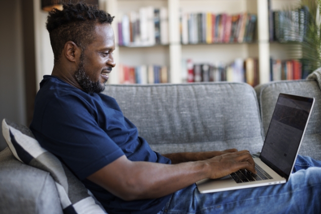 Man sits on couch typing on his laptop and smiling.