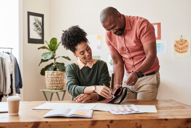 Two professionals collaborating on a tablet at a wooden desk in a modern home office, with business documents and charts spread out on the table
