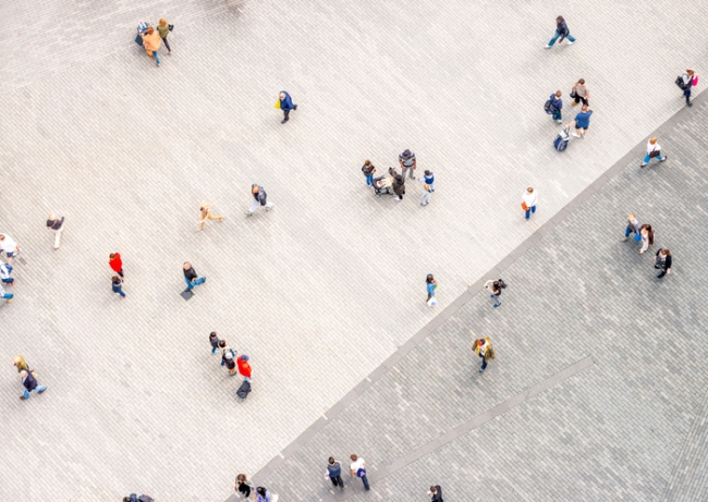 Aerial view of people walking on a large, light-colored paved plaza divided by a diagonal line into two slightly different shades of gray paving stones.