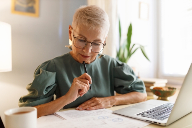 Concentrated woman with short hair and glasses reading documents at her desk with a pen and laptop.