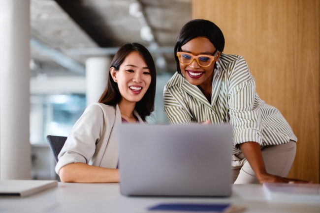 Two women smiling and looking together at a laptop screen in an office setting.