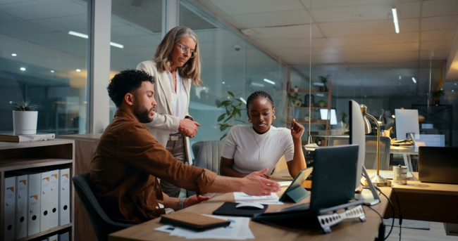 Three colleagues, two sitting and one standing, working together around a computer screen in a modern office.
