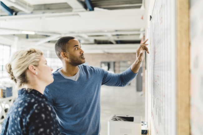 A man is pointing at a whiteboard while a woman tilts her head and looks at what he's pointing at.