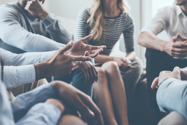  Close-up of a group of people sitting in a circle, with a man's gesturing hands prominent in the foreground.