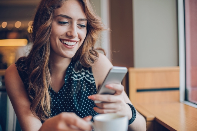 Smiling woman in polka dot top looking at smartphone with coffee cup at cafe table