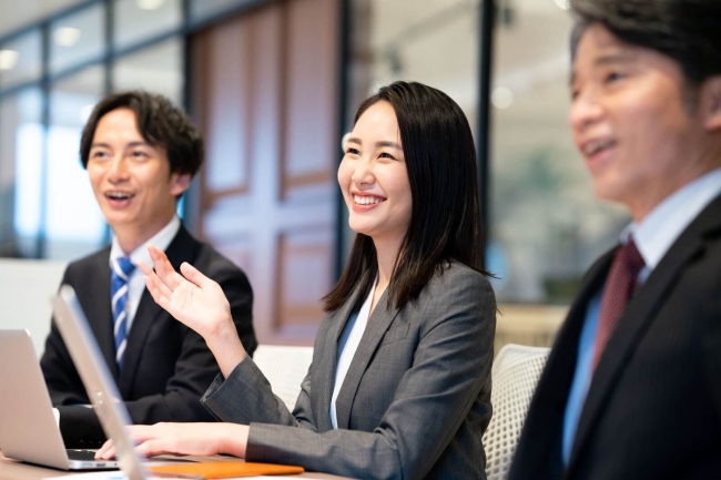 会議中に笑顔で発言するスーツ姿の女性と、両隣に座る2人の男性同僚。