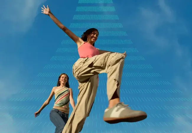 A low-angle shot of two women leaping into a blue sky, with a graphic pattern of ascending lines creating perspective.