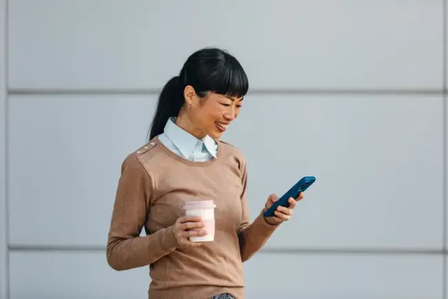 コーヒーカップを持ちながら屋外でスマートフォンを確認する笑顔の女性。