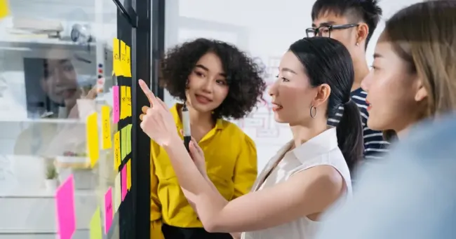 ブレインストーミング中にガラスボードに貼られたカラフルな付箋を確認するチームメンバーたち。
