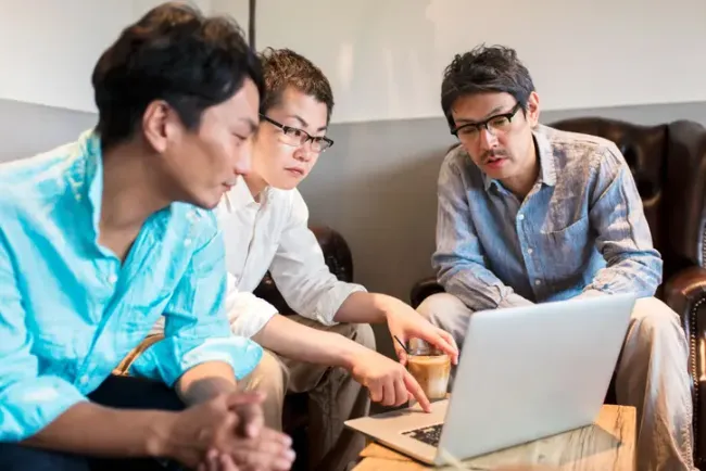 カジュアルな打ち合わせスペースでノートパソコンの画面を見ながら話し合う3人の同僚。