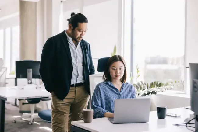 モダンなオフィスでノートパソコンに向かう女性の横に立ち、画面を一緒に確認する男性。
