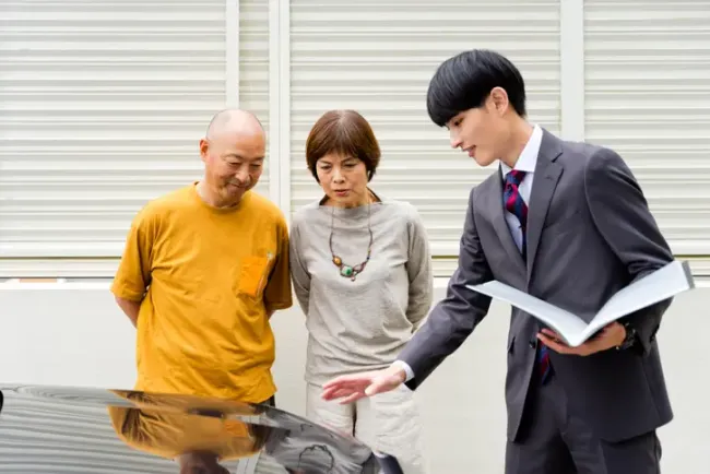 車のそばでカタログを見せながら夫婦に説明する自動車販売員。