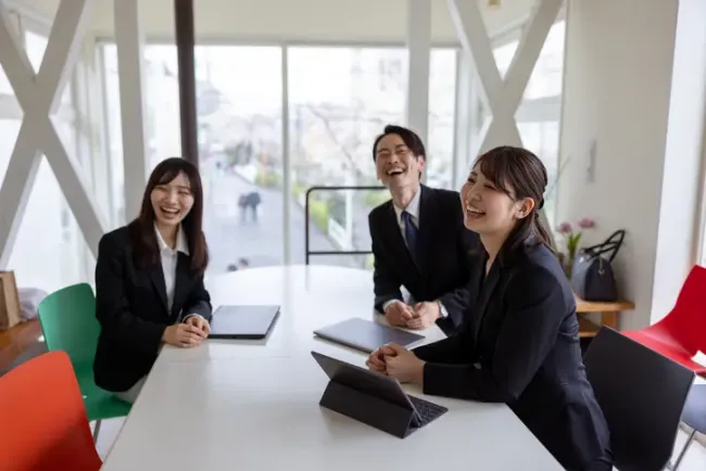 タブレットやノートパソコンを前に会議テーブルで笑い合うビジネススーツ姿の3人の同僚。