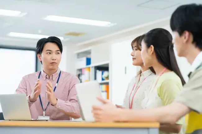 ネームタグを付けた若手社員が、同僚たちに身振りを交えて話しかけている様子。