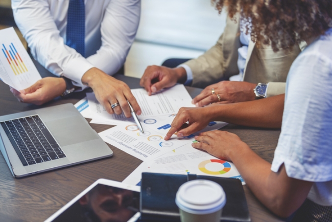 A group of colleagues sitting around a table review and point to various printed market research statistics, charts, and data visualizations.