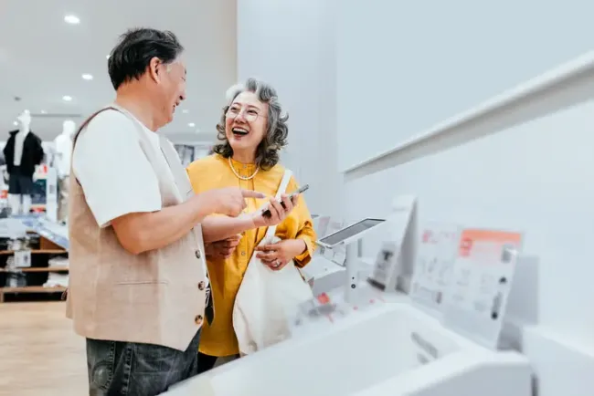 家電量販店のカウンターでスマートフォンを比較しながら笑い合う年配のカップル。