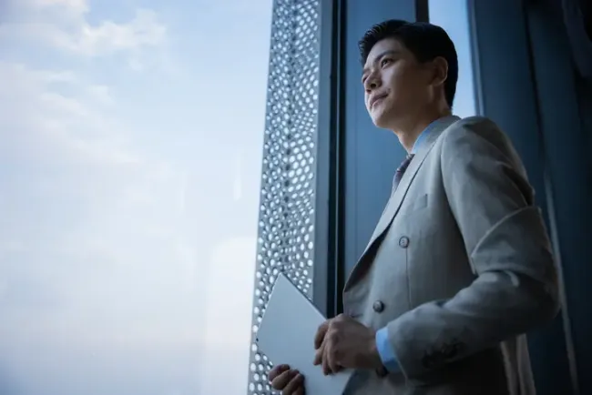 高層ビルの窓から外を見つめる、ノートパソコンを抱えたスーツ姿のプロフェッショナル。