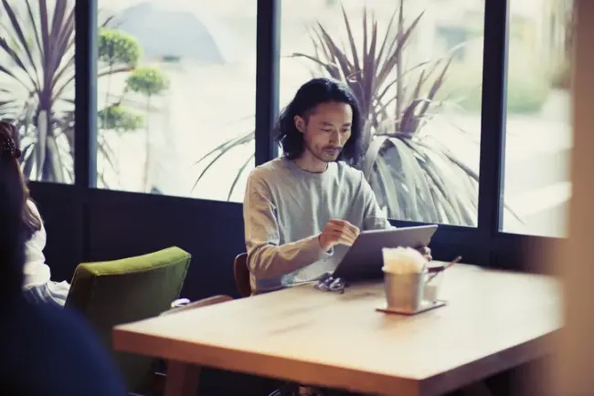 植物が見える窓際のカフェテーブルでタブレットを読む男性。