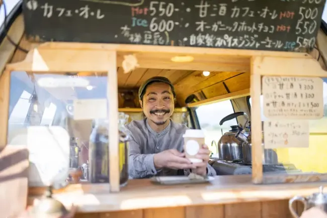 手書きの日本語メニューボードがあるフードトラックからコーヒーを提供する笑顔の店主。