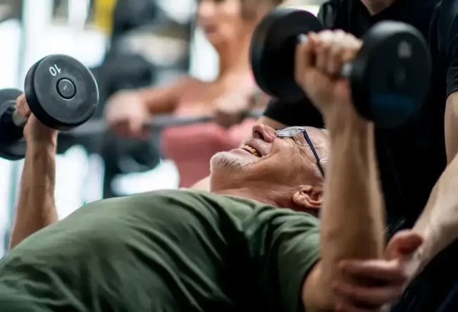 Man bench pressing dumbells with assistance from personal trainer.