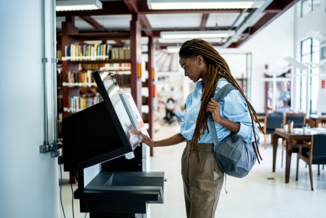  A young woman with a backpack uses a touchscreen kiosk in a modern library or bookstore.