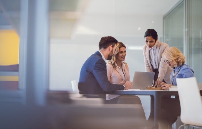 A stock image of several professionals gathered around a table looking at a laptop