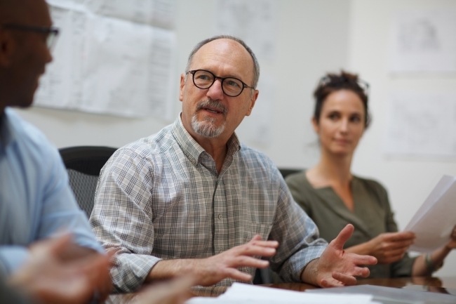 A stock image of a mid-age white man speaking at a meeting with others.