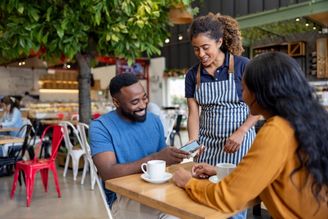 Waitress taking payment and laughing with customers at coffee shop.