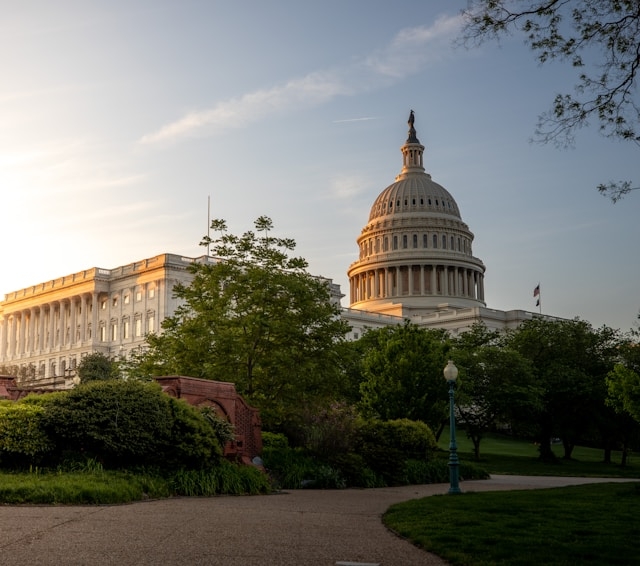The U.S. Capitol Building dome and a nearby white government building at sunset, viewed through green trees and a park walkway.