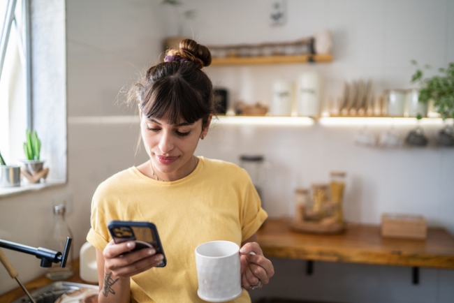 Young woman using social media on her mobile phone while drinking coffee at home.