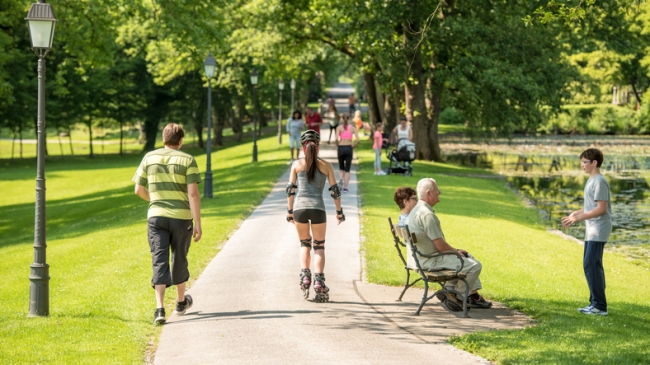 People walking on footpath, senior couple sitting on bench, young woman rollerblading in park.
