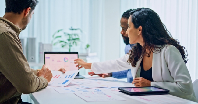 Three business professionals in a meeting reviewing customer intelligence data and charts on a laptop screen and printed documents.