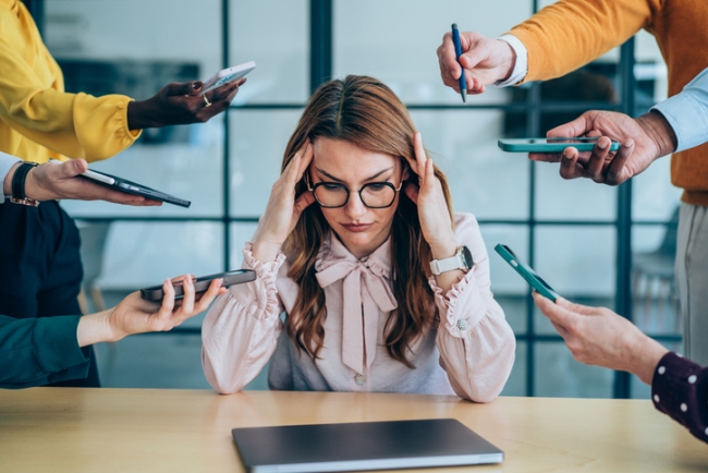 Stressed out female employee surrounded by colleagues asking her for feedback.