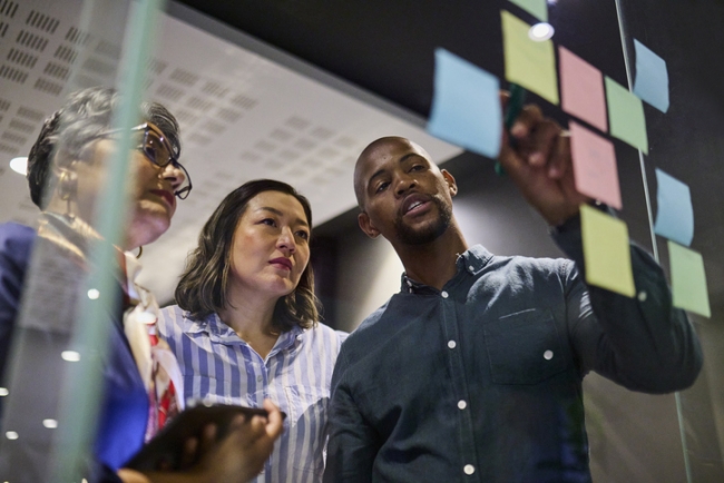 Professionals collaborate on a glass board covered in colorful sticky notes, discussing ideas and strategizing.