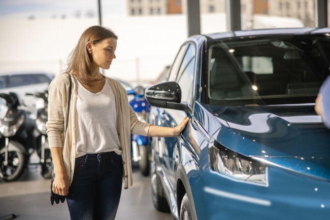 Woman looking for a new car, standing next to a new SUV indoors at a car dealer, touching the door.