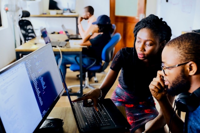 Two students, a man and a woman, collaborate while looking at a computer screen in a modern classroom.