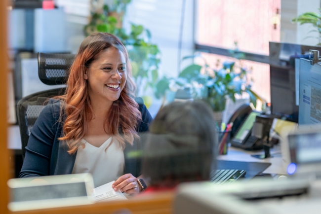 View through an office window of a female executive sitting at her desk and smiling while talking to employees..