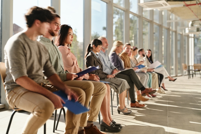 Large number of candidates sitting in the corridor by a window waiting for a job interview.