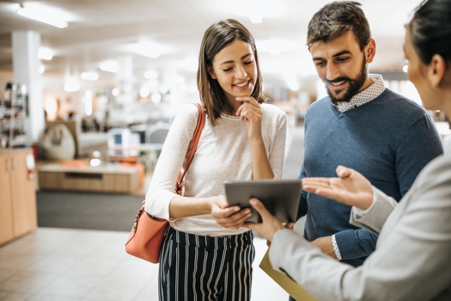 A saleswoman using a digital tablet to assist a smiling young couple in a modern retail showroom.