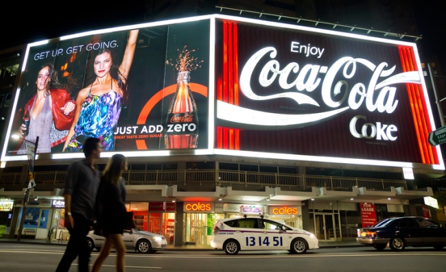 A young couple look at two large neon Coca-Cola billboards as they walk past traffic on Kings Cross at night. 